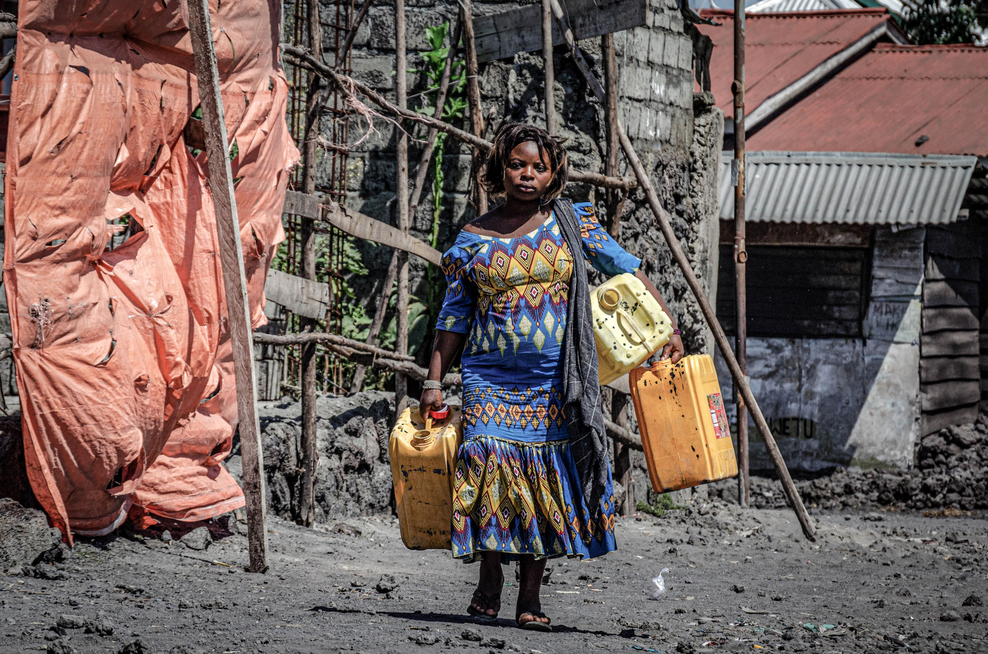 Goma, RDC, mai 2020. Une femme transporte des jerricans d’eau dans une rue du chef-lieu du Nord-Kivu. Au début de l’épidémie de coronavirus, la régie publique Regideso a promis un accès gratuit à l’eau, mais vu le peu de gens raccordés au réseau, par ailleurs très défectueux, le manque d’eau a entravé le respect des normes sanitaires et des mesures d’hygiène et a suscité de nombreuses manifestations dans la ville, étalée sur les rives du lac Kivu. © Arlette Bashizi pour la Fondation Carmignac