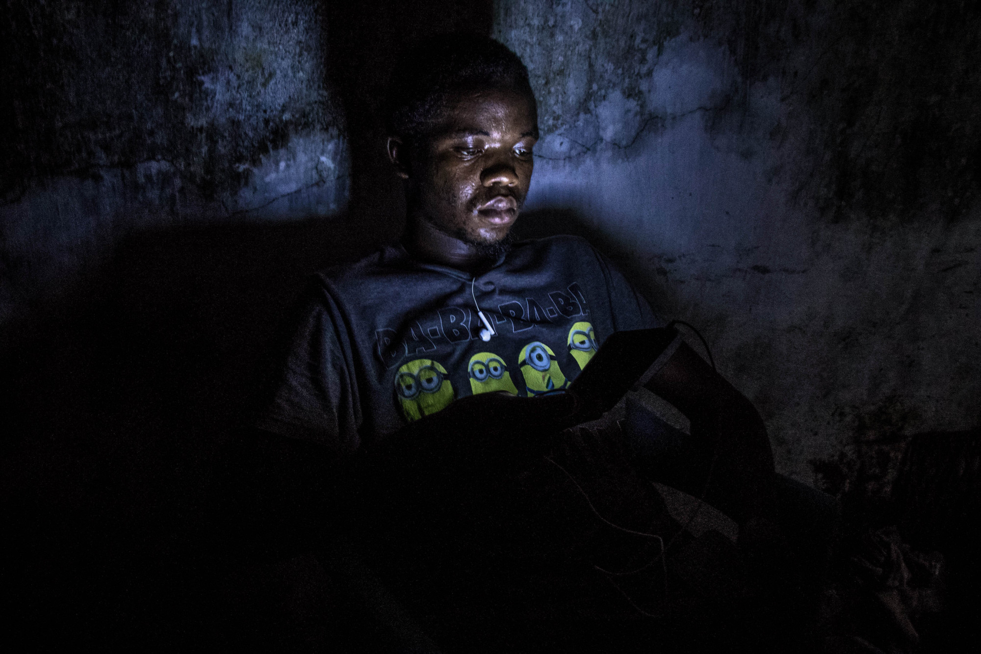 Mbudi, Kinshasa, RDC, mai 2020. Après avoir rechargé son téléphone contre paiement à une borne communale alimentée par générateur, le jeune David lit sur son écran dans les ténèbres. © Justin Makangara pour la Fondation Carmignac
