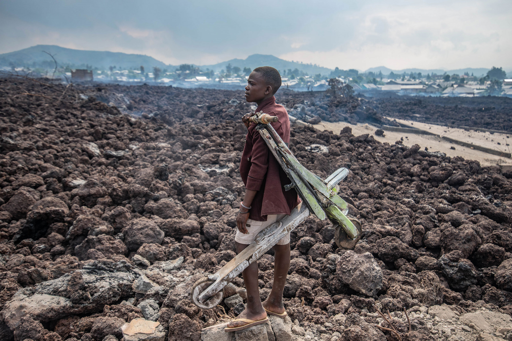 North Kivu, 23 May 2021. A boy carrying a Chukudu, or wooden bike, surveys lava cooling in the Buhene neighborhood after the eruption of Nyiragongo volcano. © Moses Sawasawa for Fondation Carmignac